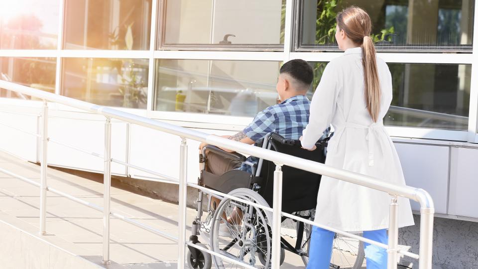 Ärztin schiebt Person im Rollstuhl eine Rampe hinauf