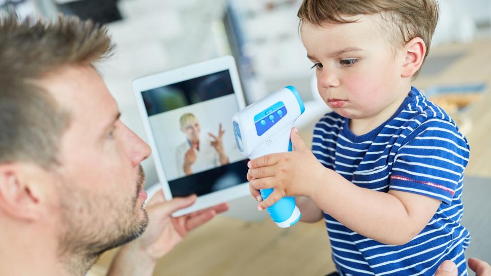 Vater sitzt mit einem Kleinkind am Tisch, der Junge spielt mit einem Fieberthermometer, der Vater hält ein Tablet mit dem Bild einer Ärztin in der Hand