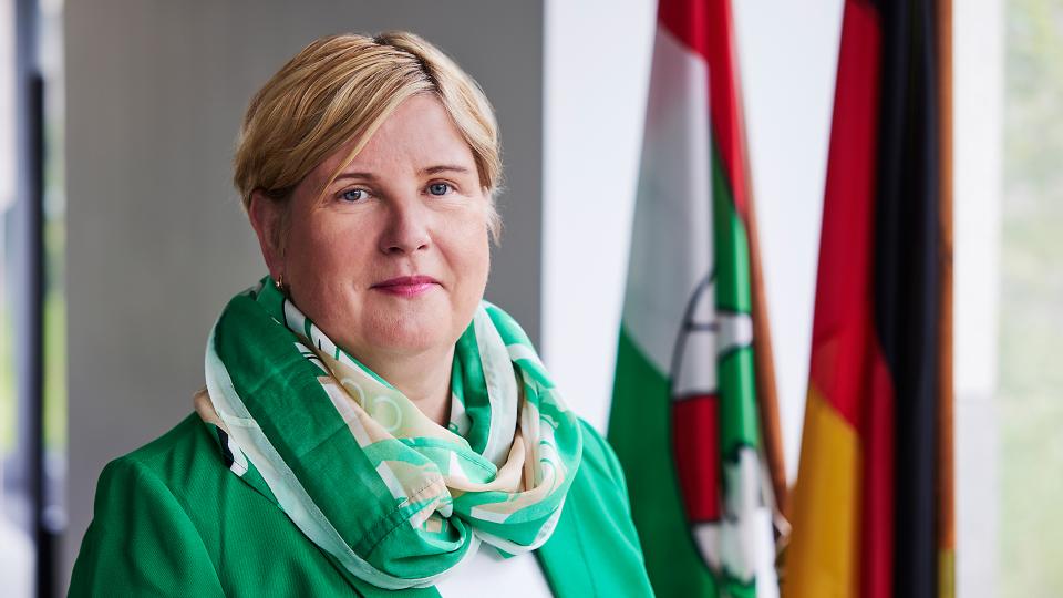 Frau Middendorf lächelt in die Kamera. Sie trägt eine weißen Bluse, einem grünen Blazer und einen farblich passenden leichten Schal. Im Hintergrund sind die Fahnen des Landes Nordrhein-Westfalens und Deutschlands zu sehen.