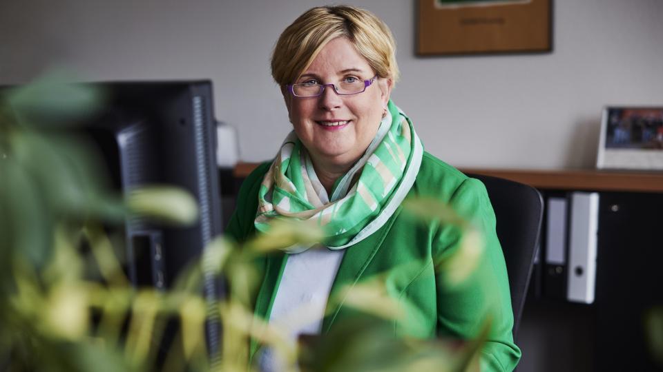 Claudia Middendorf sitzt in ihrem Büro am Schreibtisch am PC und lächelt in die Kamera. Sie trägt eine weiße Bluse, einen grünen Blazer und einen farblich passenden Schal.
