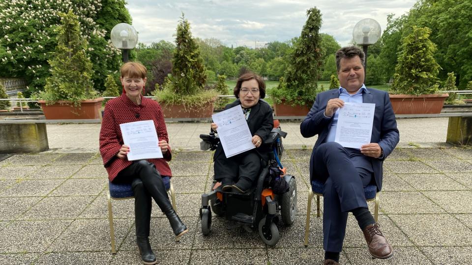 Prof Dr. Helga Seel, Rika Esser und Jürgen Dusel (von links nach rechts) sitzen nebeneinander auf einer Terasse mit Steinplatten. Alle halten die Bad Nauheimer Erklärung in den Händen und in die Kamera.