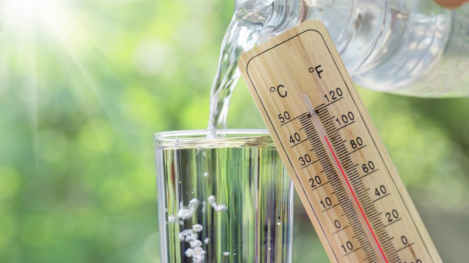 In ein Glas wird Wasser eingeschenkt. Rechts daneben wird ein Thermometer ins Bild gehalten auf dem 40 Grad angezeigt werden. Im Hintergrund sieht man verschwommen Sonnenstrahlen, die durch grüne Bäume scheinen.