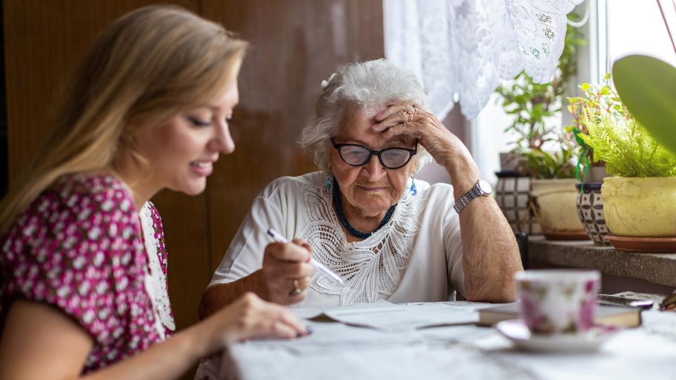 Am linken Bildrand sitzt eine junge blonde Frau zusammen mit einer älteren Frau (Bildmitte) an einem Tisch am Fenster. auf dem Tisch liegt eine weiße Tischdecke. Darauf steht eine weiße Kaffeetasse mit Blumenmuster in rosa. Auf der Fensterbank am rechten Bildrand stehen einige Grünpflanzen. 