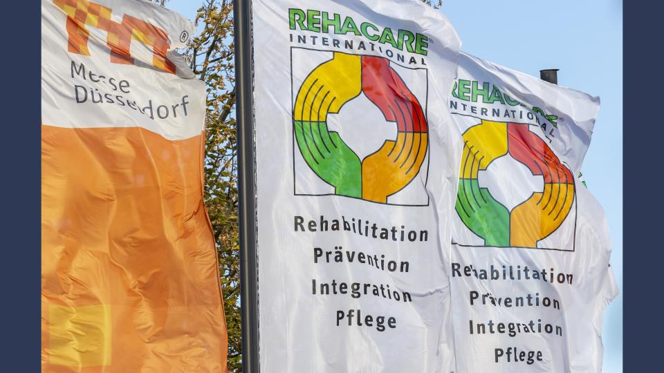 Im Freien stehen drei Fahnen nebeneinander. Die erste am linken Bildrand ist orange mit dem Logo der Messe Düsseldorf. Die beiden daneben sind weiß und tragen das Logo und die Schlagworte der Rehacare.