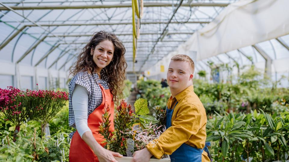 Ein junger Mann mit Down Syndrom und eine Frau stehen in einer Gärtnerei und halten gemeinsam eine Kiste Blumen zwischen sich.