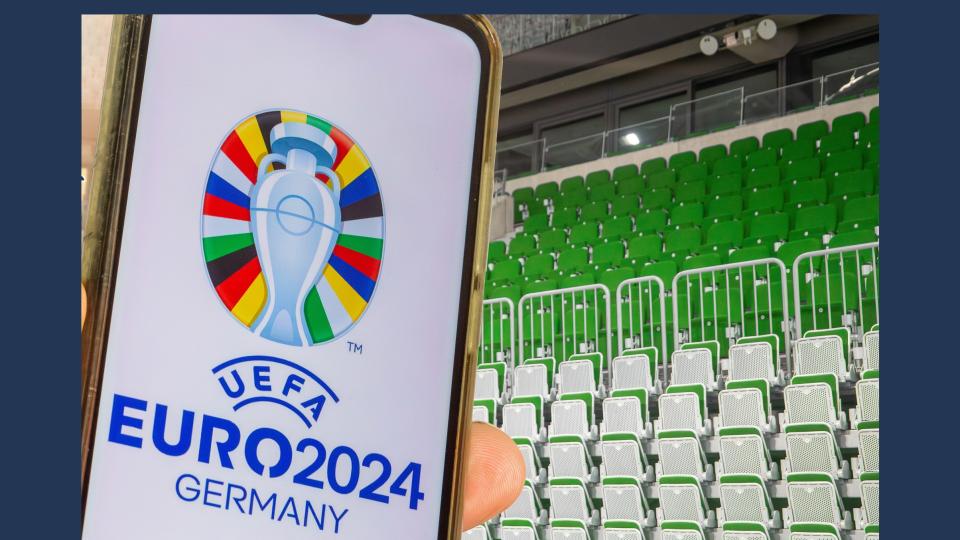 Auf dem Bild befinden sich im Hintergrund grüne Sitzplätze. Am linken Bildrand wird ein Handy in der Hand gehalten. Auf dem Display ist das Logo der Europameisterschaft abgebildet, ein Pokal auf unterschiedlichen Flaggen mit dem Schriftzug "UEFA EURO2024 GERMANY" darunter.