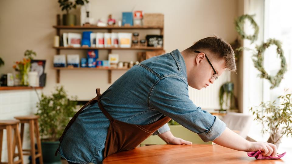 Ein junger Mann mit Down-Syndrom arbeitet in einem Café. Er trägt ein blaues Hemd und eine braune Schürze und wischt gerade einen Holztisch ab. Im Hintergrund steht ein Teil der Theke und es hängt ein Regal an der Wand. Darauf steht eine Pflanze und verschiedene Packungen Kaffee.