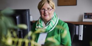 Claudia Middendorf sitzt in ihrem Büro am Schreibtisch am PC und lächelt in die Kamera. Sie trägt eine weiße Bluse, einen grünen Blazer und einen farblich passenden Schal.