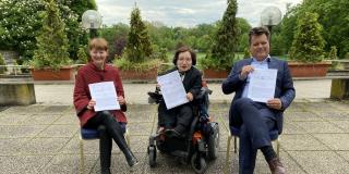 Prof Dr. Helga Seel, Rika Esser und Jürgen Dusel (von links nach rechts) sitzen nebeneinander auf einer Terasse mit Steinplatten. Alle halten die Bad Nauheimer Erklärung in den Händen und in die Kamera.