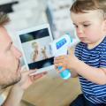 Vater sitzt mit einem Kleinkind am Tisch, der Junge spielt mit einem Fieberthermometer, der Vater hält ein Tablet mit dem Bild einer Ärztin in der Hand