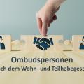 Auf einem Tischplatte aus Holz stehen drei Puzzelstücke aus Holz, die noch nicht ineinandergreifen. Auf dem Puzzleteil links ist ein Mann und auf dem rechten Puzzleteil eine Frau. In der Mitte sind schüttelnde Hände abgebildet. Darunter steht "Ombudspersonen nach dem Wohn- und Teilhabegesetz".